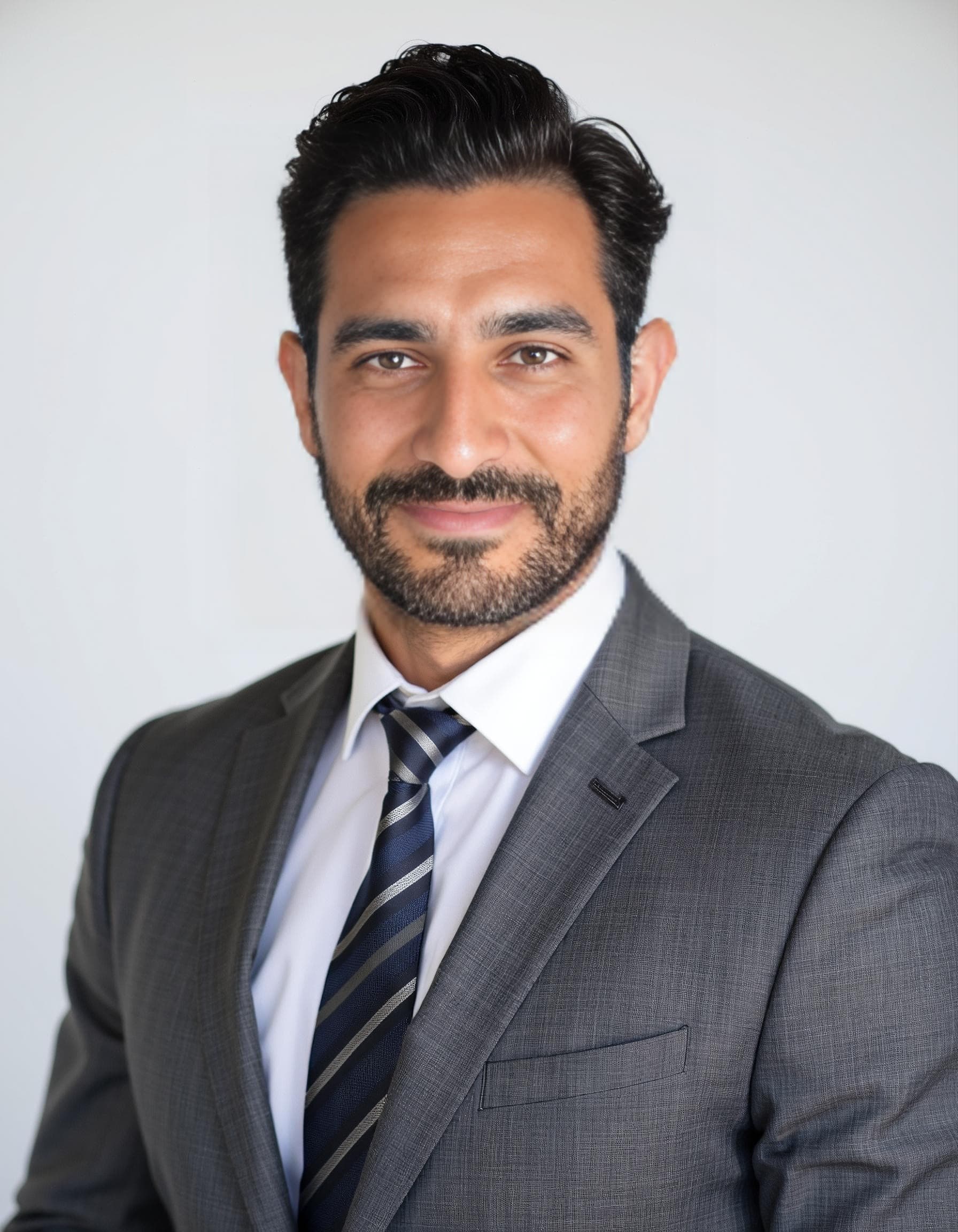 A bearded man in a charcoal suit and navy-silver striped tie smiles against a light studio backdrop in an AI headshot.