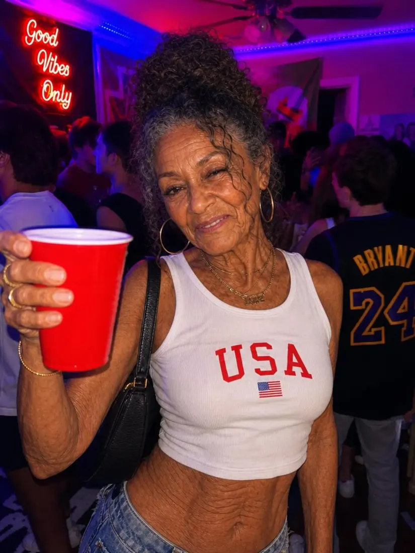 An elderly woman with curly gray hair holds a red cup at a neon-lit party, aged forward by the Aged Filter tool.