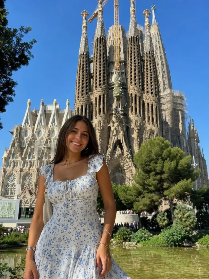 A woman in a blue floral dress stands before a crisply enhanced view of Barcelona's Sagrada Familia.