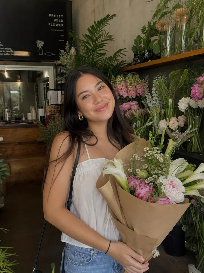 A smiling woman holds a bouquet of flowers at a flower shop, before the Block Toy Generator turns her into a brick figure.