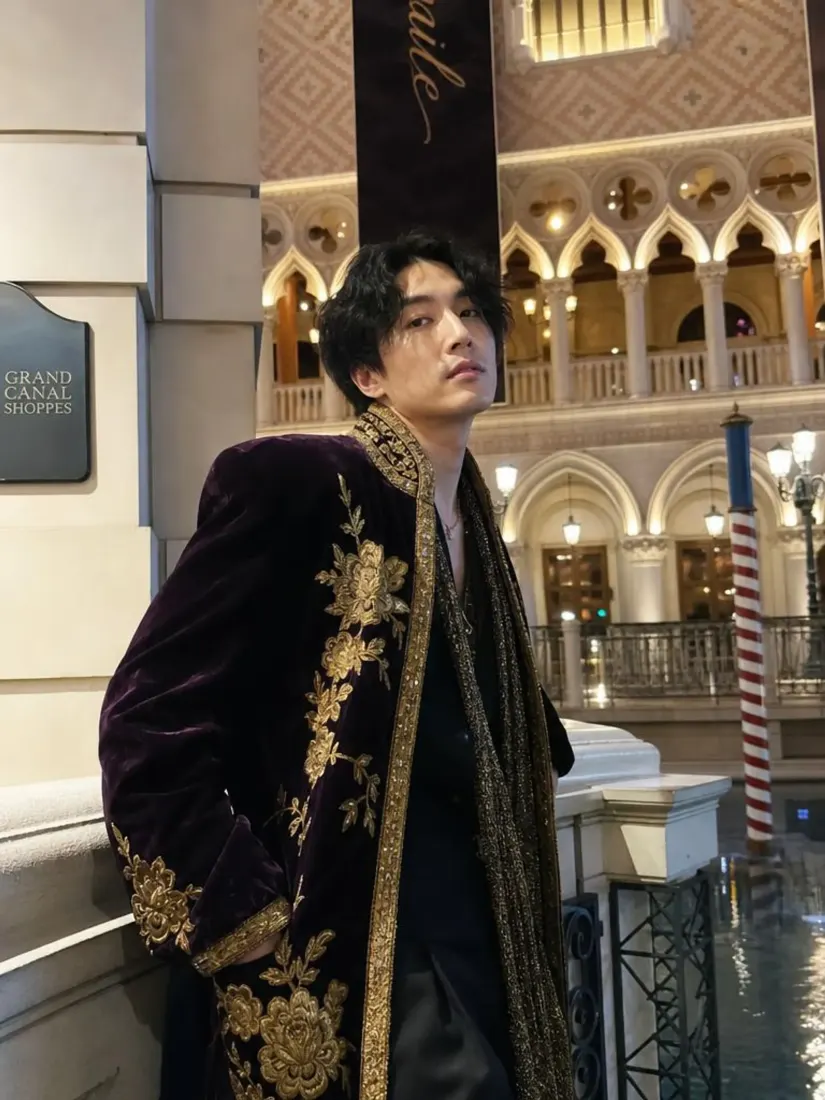 A man in a gold-embroidered purple velvet coat poses in an indoor canal shopping arcade, before the Custom Giant Doll Filter restyles him.
