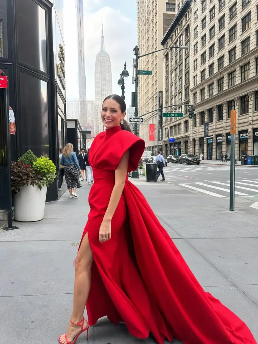 A woman in a dramatic red gown stands on a New York street with the Empire State Building visible, before the Custom Giant Doll Filter restyles her.