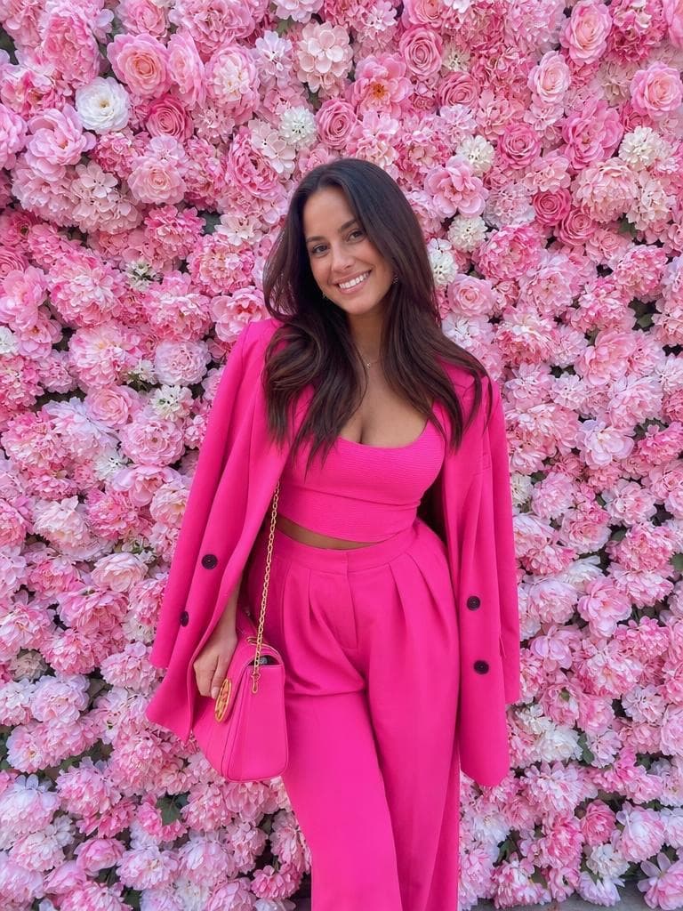 A woman in a pink suit stands against a wall of pink roses, before the Snow Globe Generator places her in a globe.