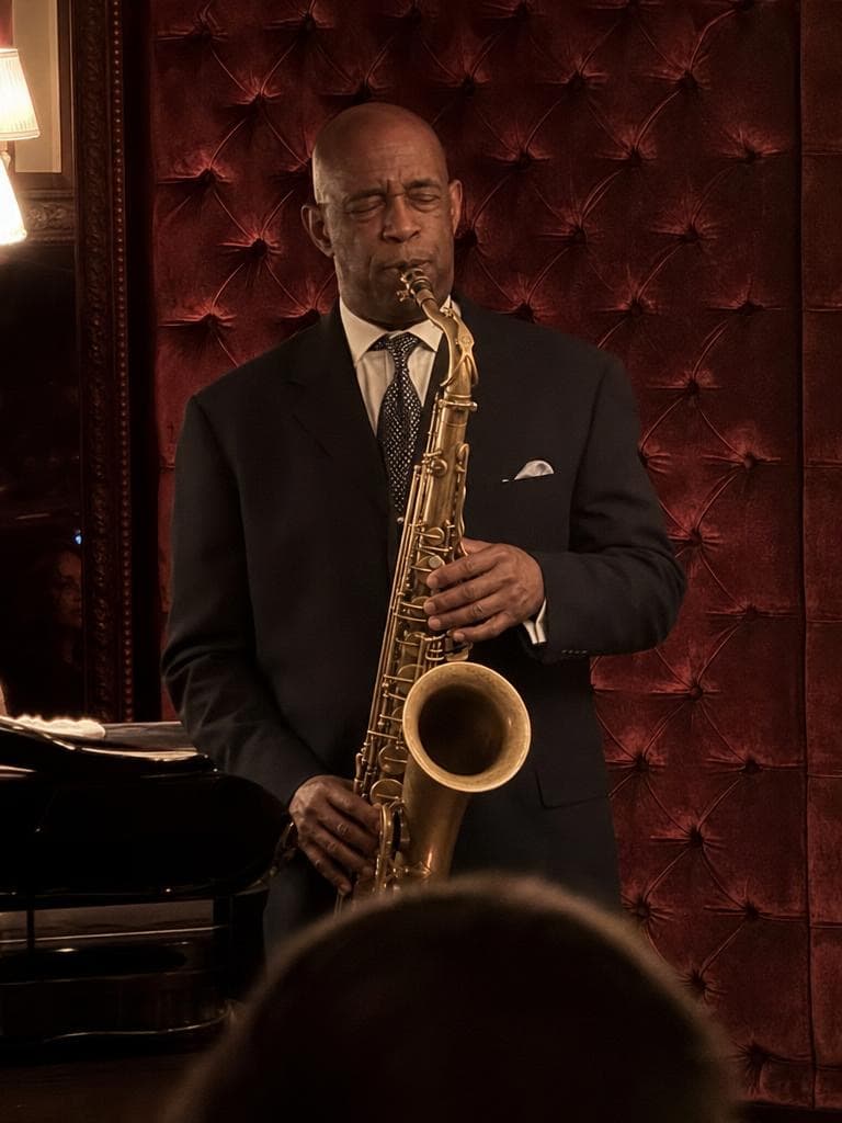 A bald man in a navy suit plays saxophone against a red tufted wall, before the Snow Globe Generator places him in a globe.