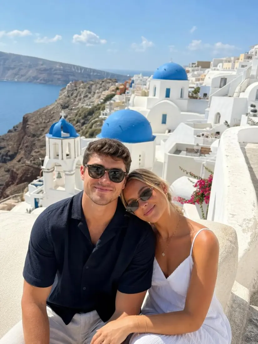 A couple in sunglasses and light clothes sit among white buildings and blue domes in daylight, before the Sunset Glow Filter warms the light.